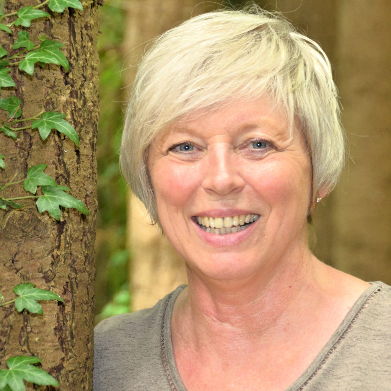 Lächelnde Frau mit kurzer, grauer Frisur, steht vor einem Baum in einem Wald.