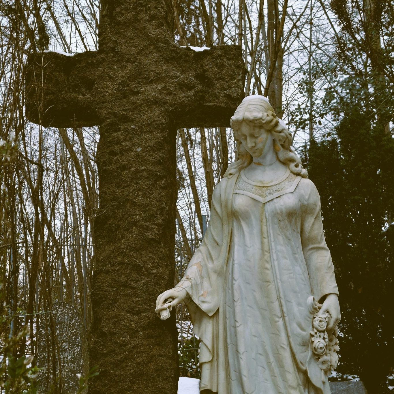 Eine Statue einer Dame mit einem Kreuz im Hintergrund, umgeben von Bäumen.