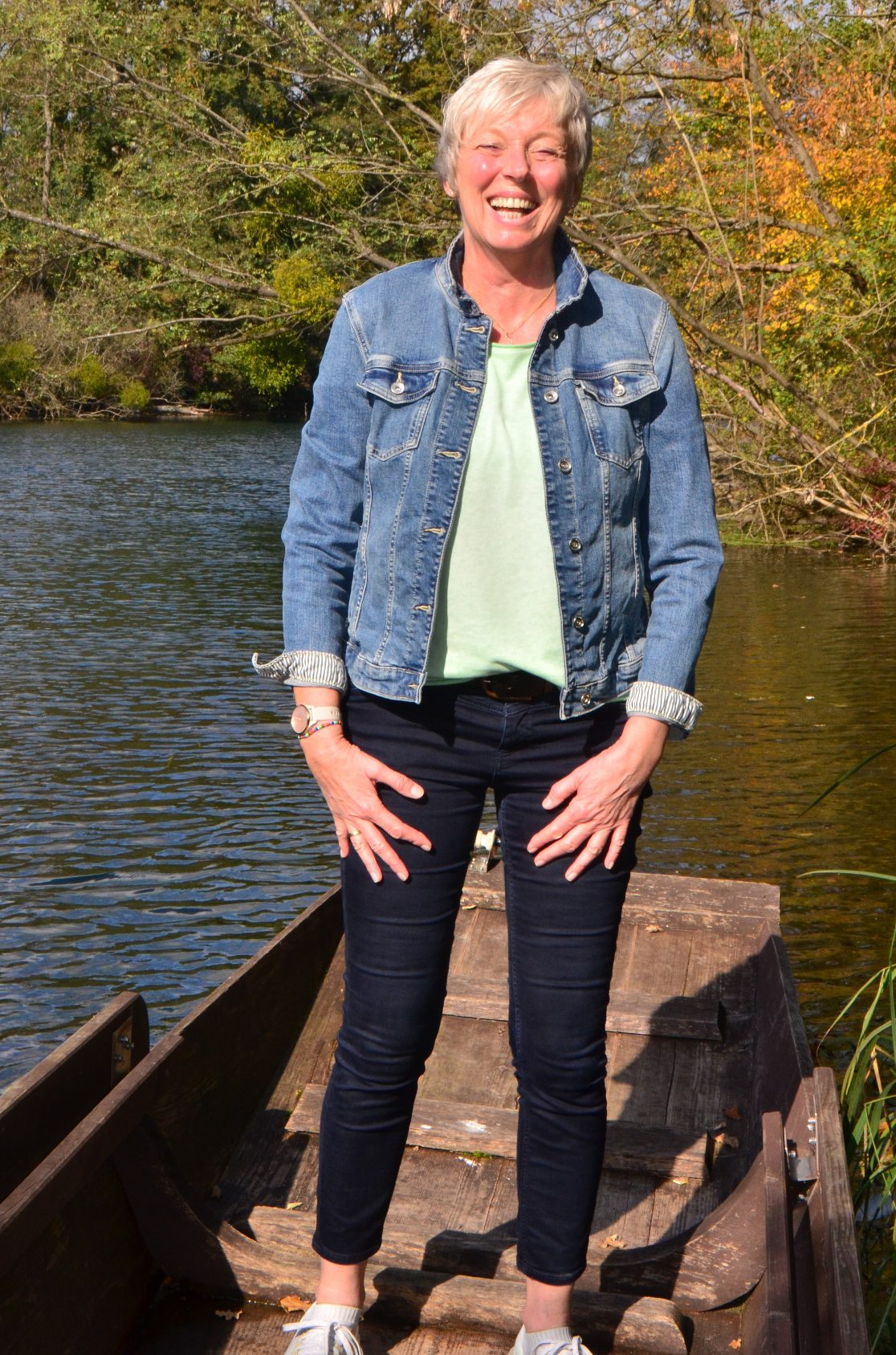 Frau steht lachend auf einem Boot vor einem Gewässer, umgeben von Bäumen.