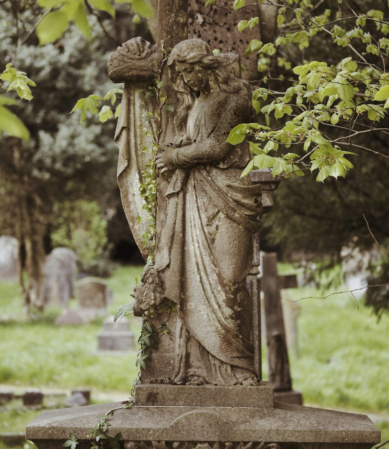 Eine Statue eines traurigen Engels mit einem Kreuz im Hintergrund, umgeben von Bäumen.